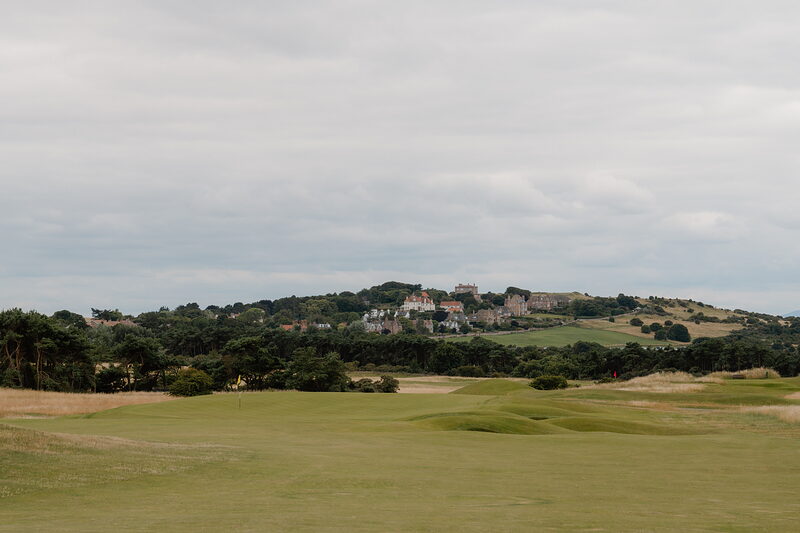 A fairway leading towards a distant clubhouse and village set among trees on a hillside