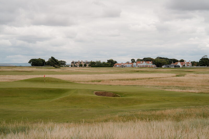 A bunker beside a green, a row of gabled hotel buildings visible across the fairway