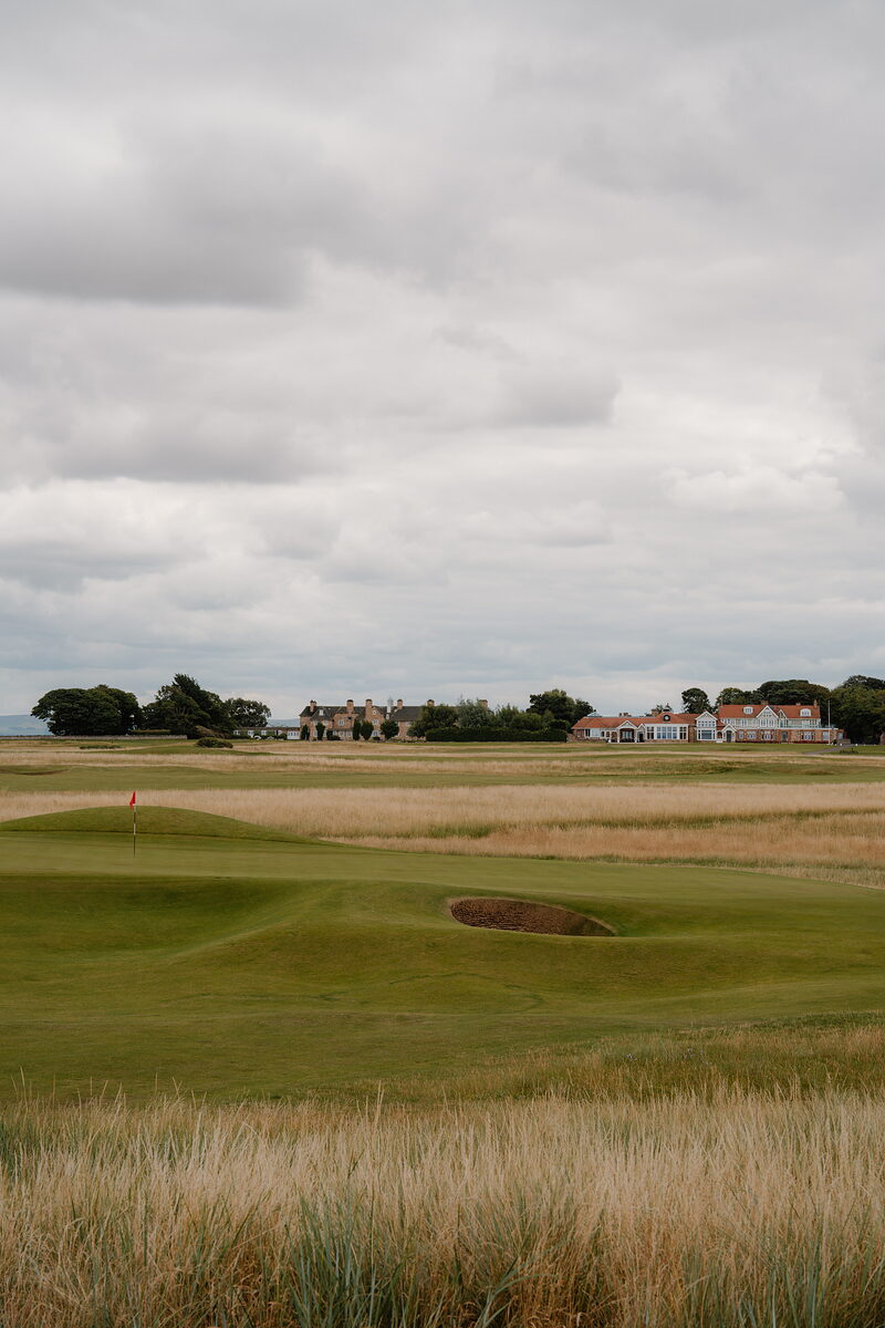 A green with a red flag and bunker set before a row of clubhouse buildings on the horizon