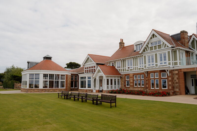 A half-timbered clubhouse with gabled bays and tall chimneys, wooden benches set on the lawn before it