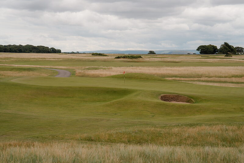 A green with a red flag set beside a bunker, a path curving through rough grass towards it