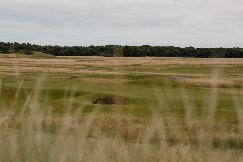 A bunker beside a green, woodland lining the fairway beyond a stretch of long grass