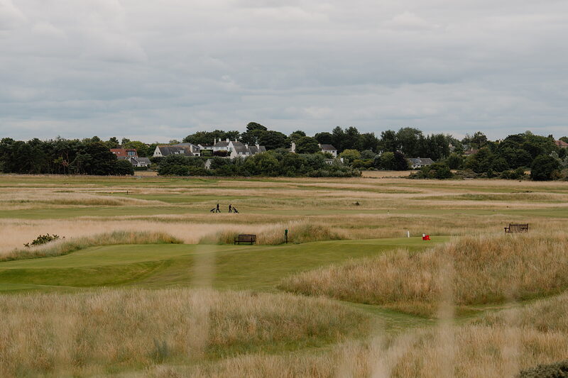 Golfers walking a fairway bordered by gorse, a town visible beyond the marshland