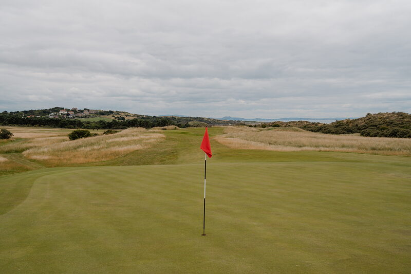 A red flag marking a green, rough grass and distant hills stretching along the horizon