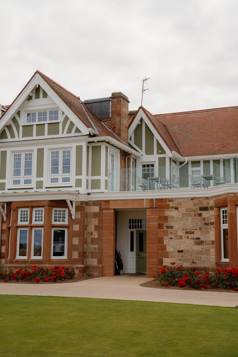 A half-timbered clubhouse with red sandstone archway and roses lining the lawn before it