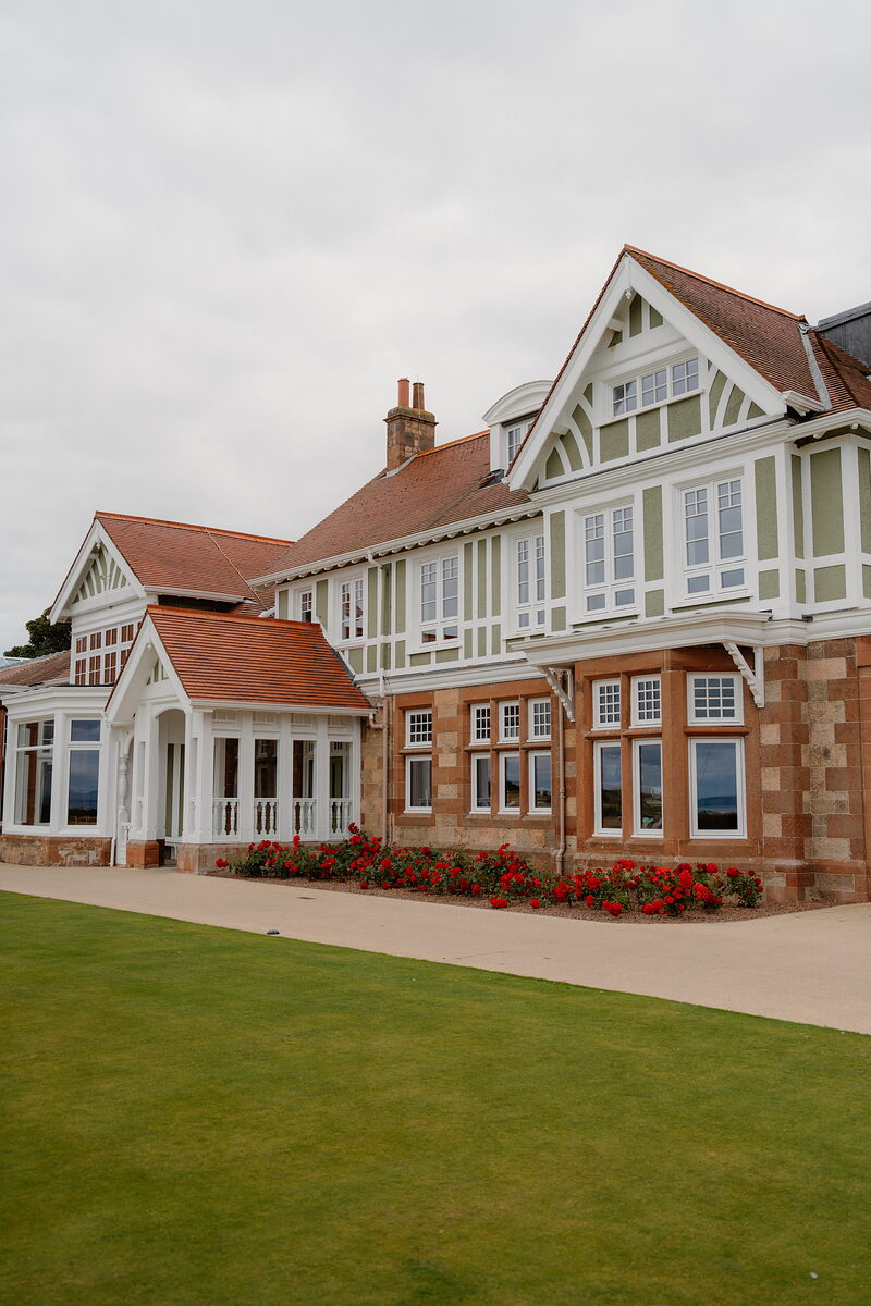 A half-timbered clubhouse with sandstone bay windows, red roses lining the path before it