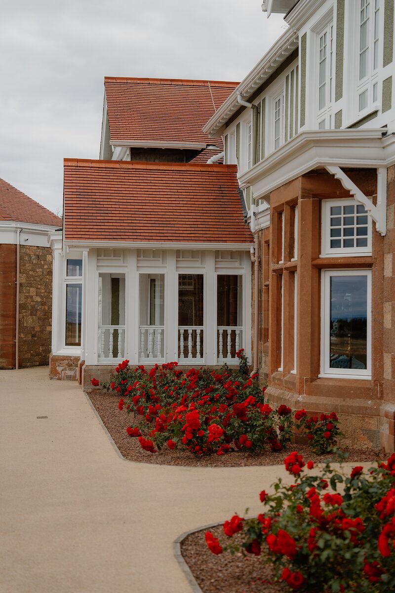 A half-timbered clubhouse wing with a covered balcony and red roses lining the path below
