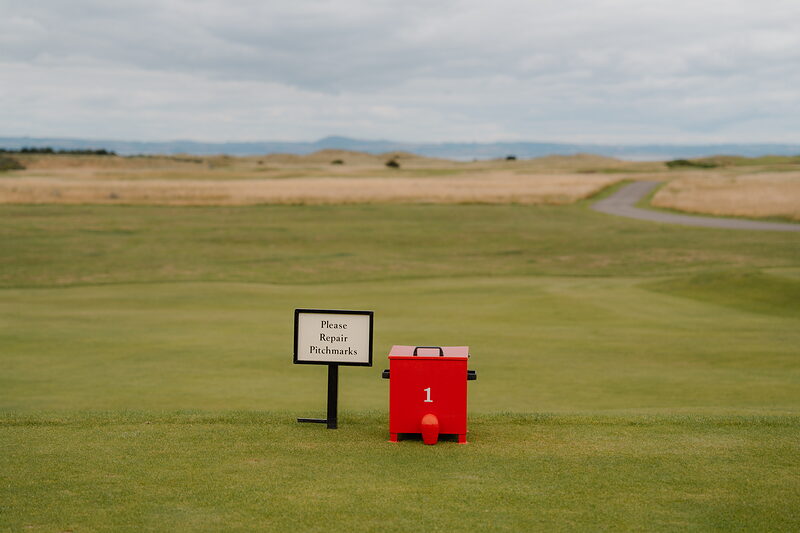 A red pitchmark repair box beside a sign reading Please Repair Pitchmarks, set on a green