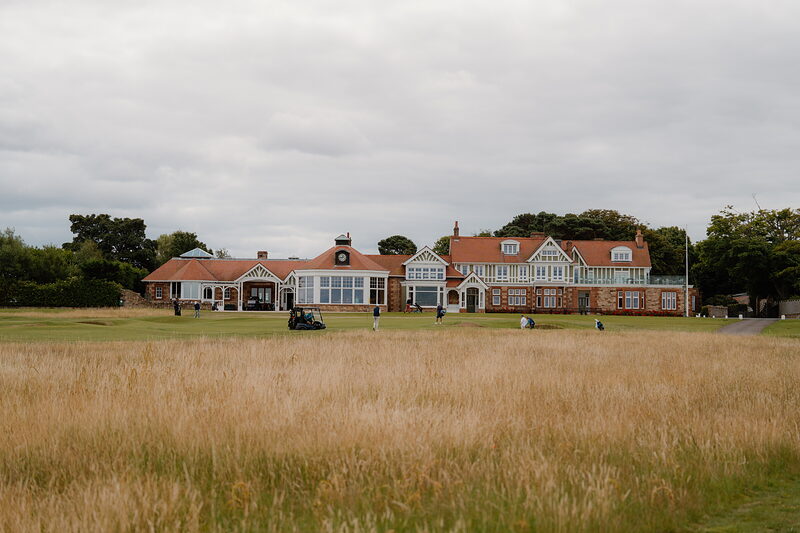 A sandstone clubhouse with tiered gables and chimneys, small figures gathered near the entrance