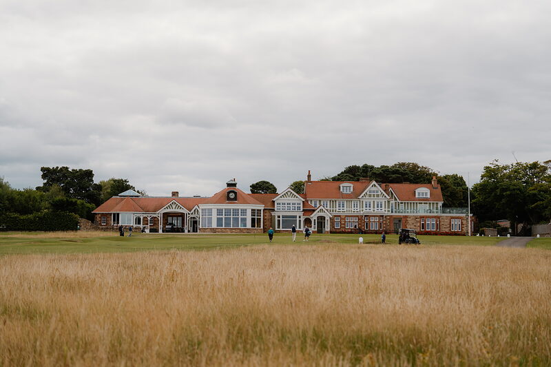A half-timbered clubhouse with sandstone chimneys, small figures gathered on the fairway before it