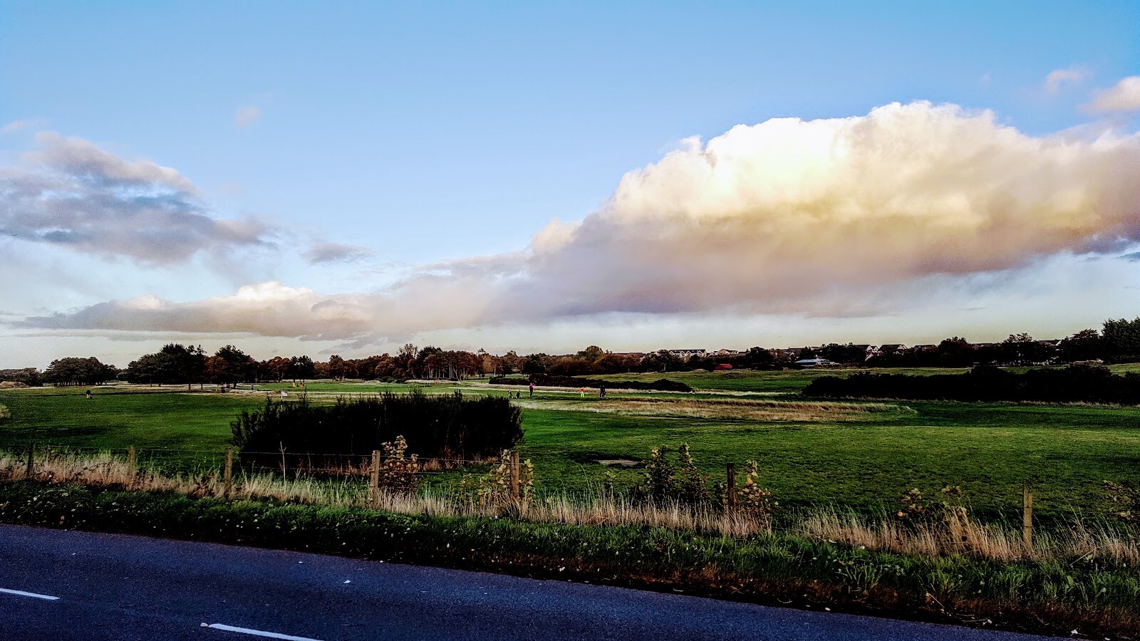 Fairways seen over a fence from the roadside, with golfers in the distance, trees and houses beyond and a big white cloud.