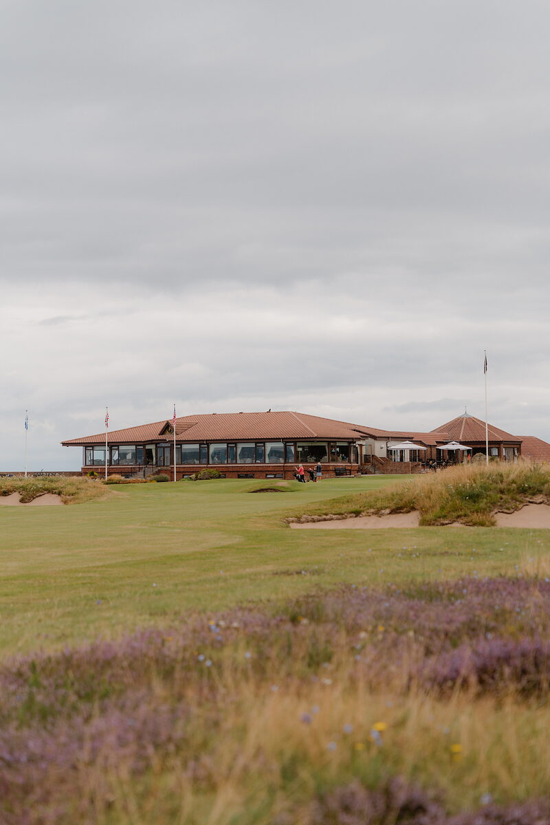 A clubhouse with a curved red roof set among bunkers and flowering heather, golfers gathered outside