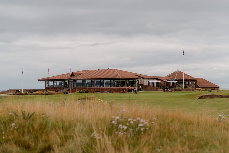 A clubhouse with a curved red roof and flagpoles set beyond a grassy fairway bordered by wildflowers