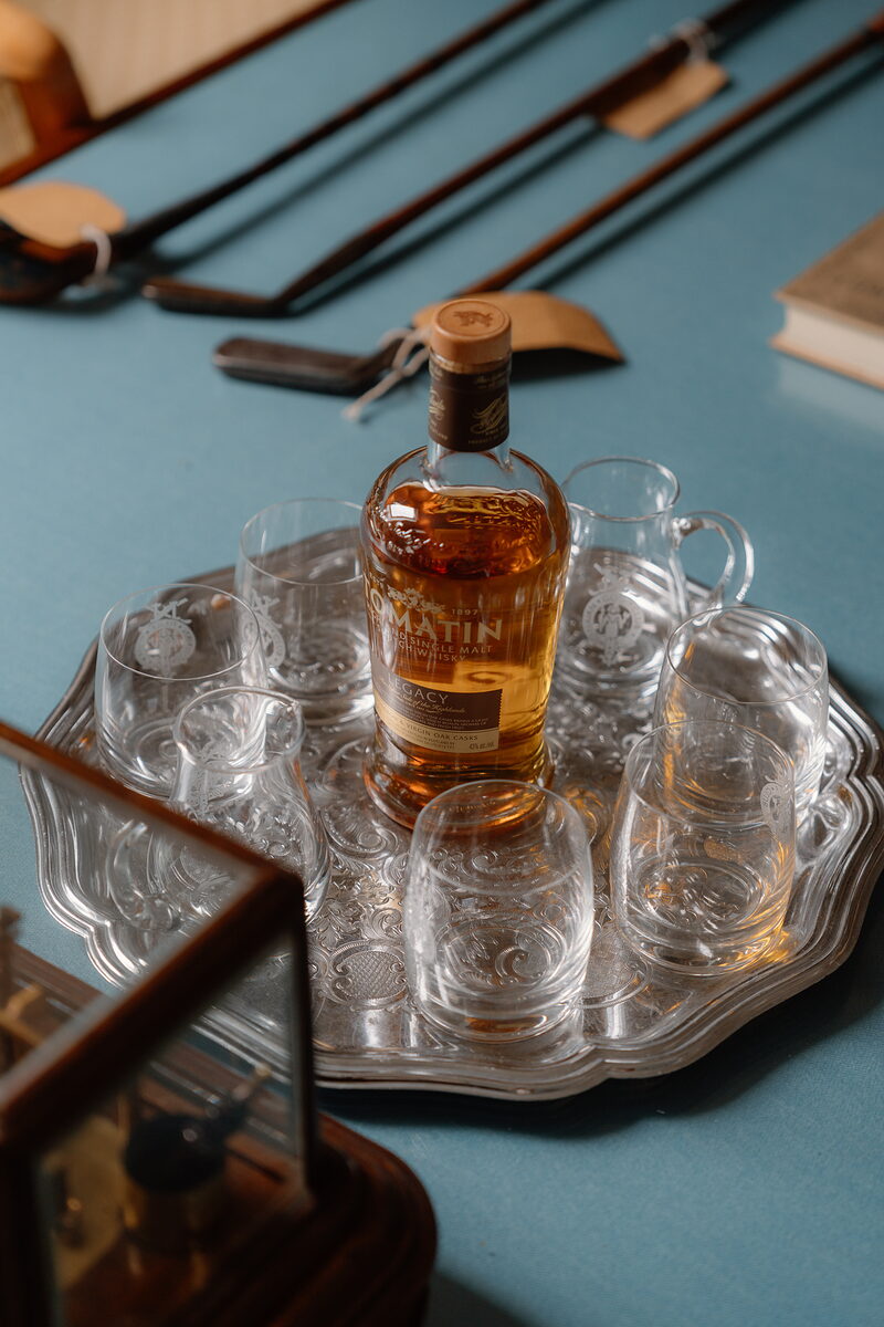 A bottle of whisky surrounded by empty glasses on a silver tray, golf clubs and a book laid out on the table behind