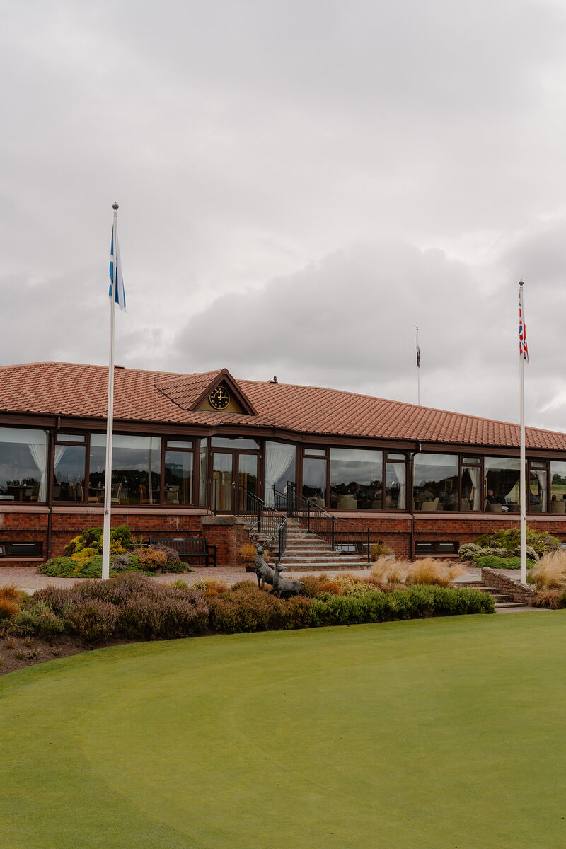 A clubhouse with a curved red roof and flagpoles set beyond a landscaped green, a stag statue among the shrubs