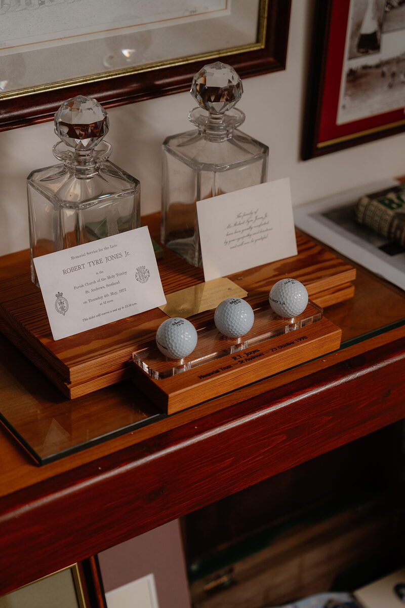 Crystal decanters and golf balls displayed on a wooden stand beside framed invitations and photographs