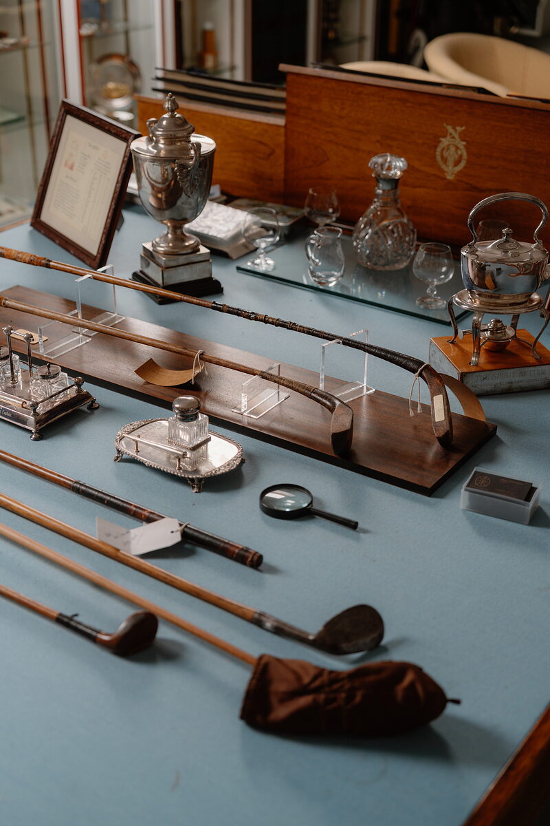 Antique golf clubs, decanters and a silver trophy displayed on a table beside a wooden presentation case