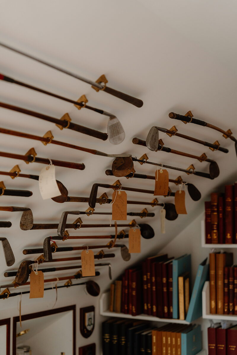 Rows of antique golf clubs with luggage tags hanging from a sloped ceiling above shelves of bound books