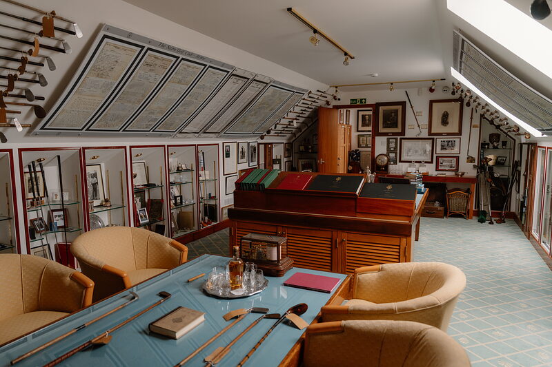 A clubhouse room lined with glass display cabinets of trophies, a chesterfield table and armchairs at its centre