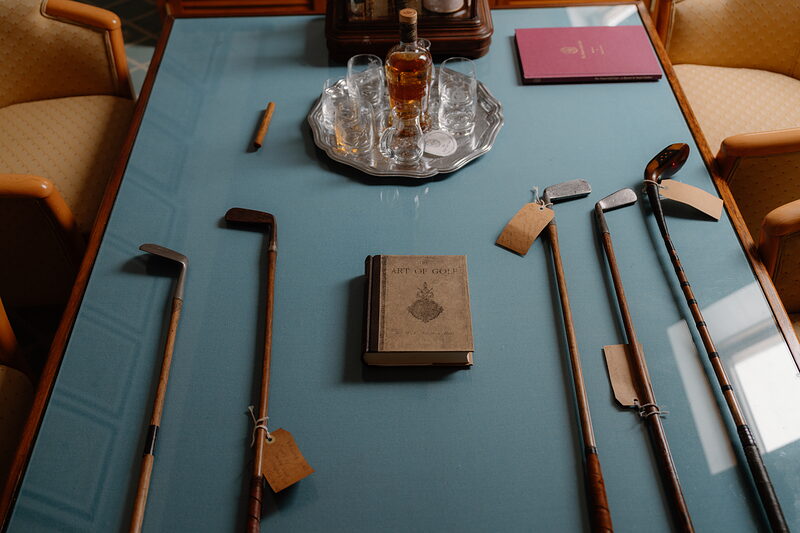 Antique golf clubs and a leather-bound book displayed on a table beside a decanter and glasses