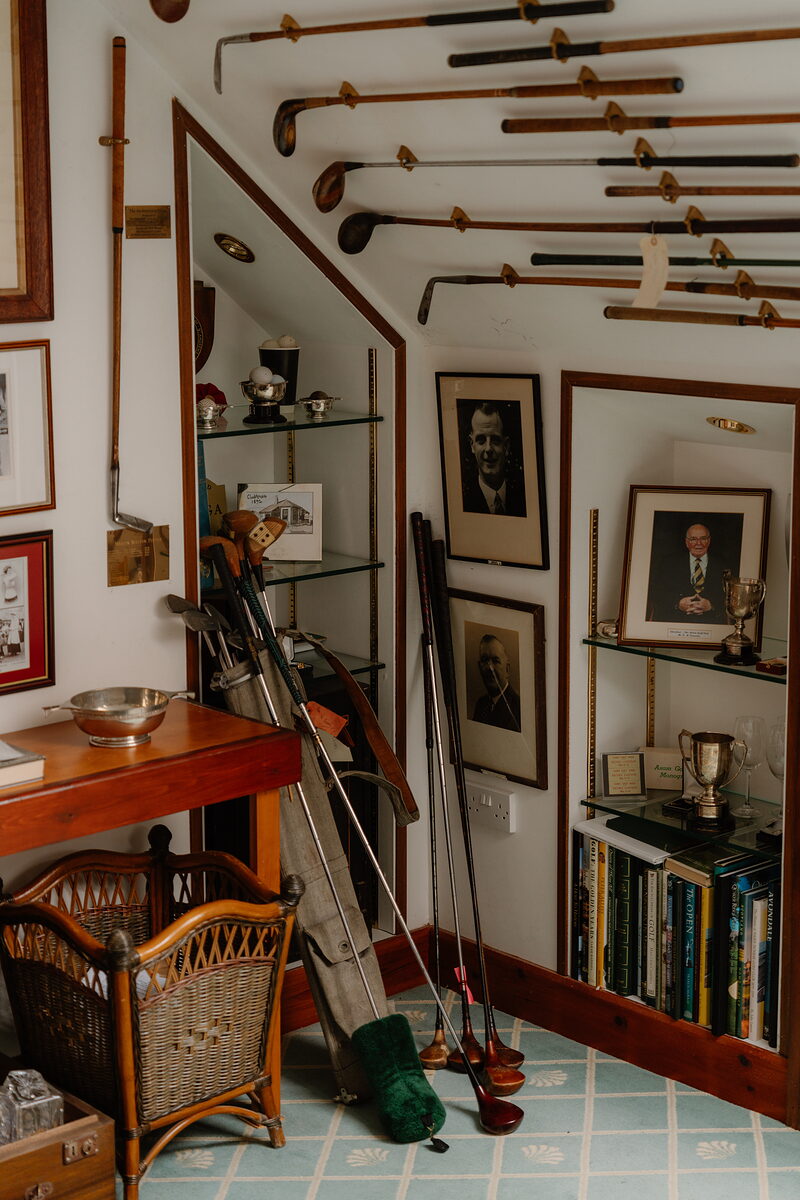 Golf clubs mounted along a sloped ceiling above framed portraits, trophies and bookshelves in a clubhouse corner