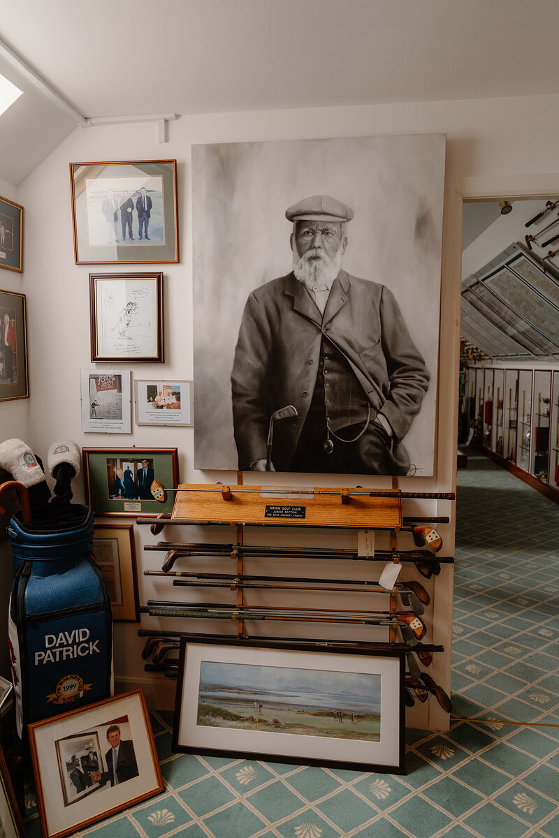 A large portrait of a bearded golfer hanging beside a rack of antique clubs and framed photographs
