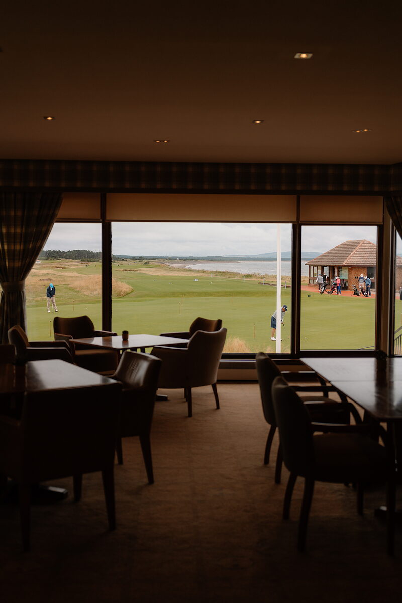 A clubhouse lounge with armchairs and tables looking out over a fairway to a distant pavilion and golfers