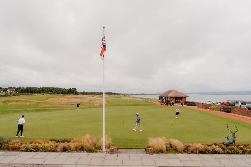 Golfers putting on a green beside a flagpole, a thatched pavilion and a bronze deer statue nearby