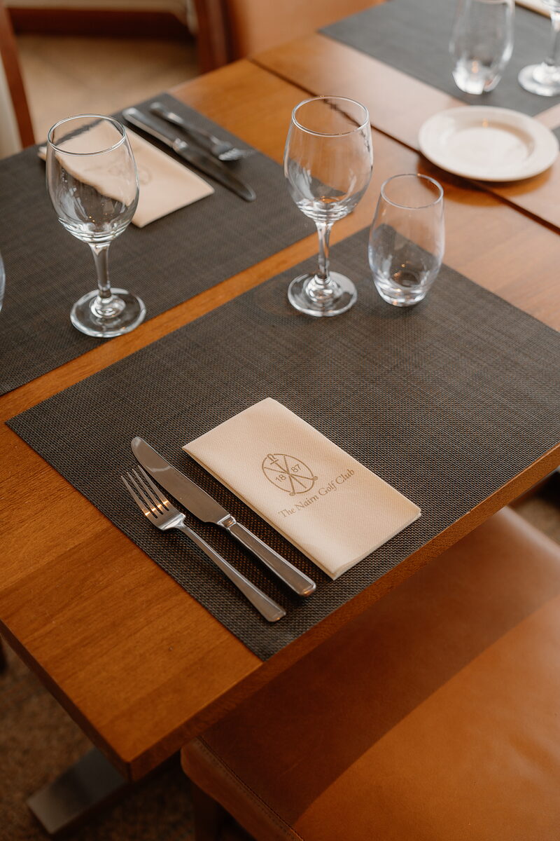 A table set with wine glasses and cutlery, a napkin embroidered with the Nairn Golf Club crest