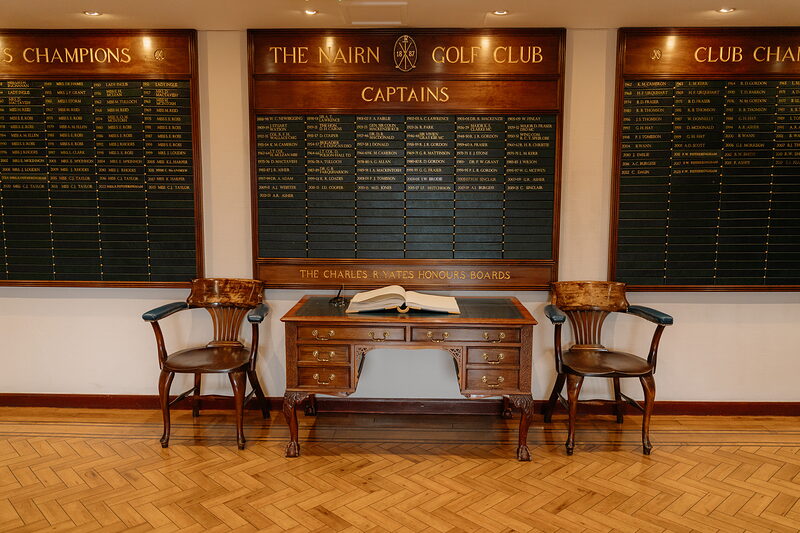 A wooden desk and chairs set beneath honours boards listing Nairn Golf Club captains and champions
