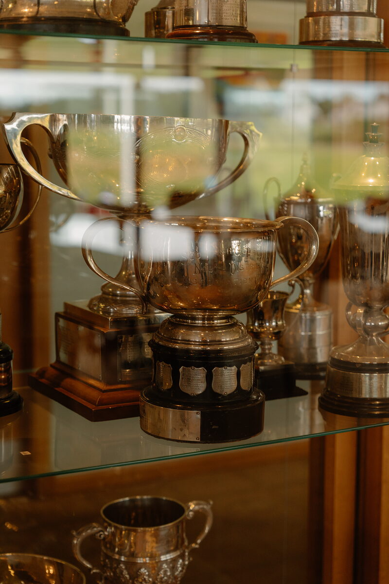 Silver trophies and cups displayed behind glass shelving in a clubhouse cabinet