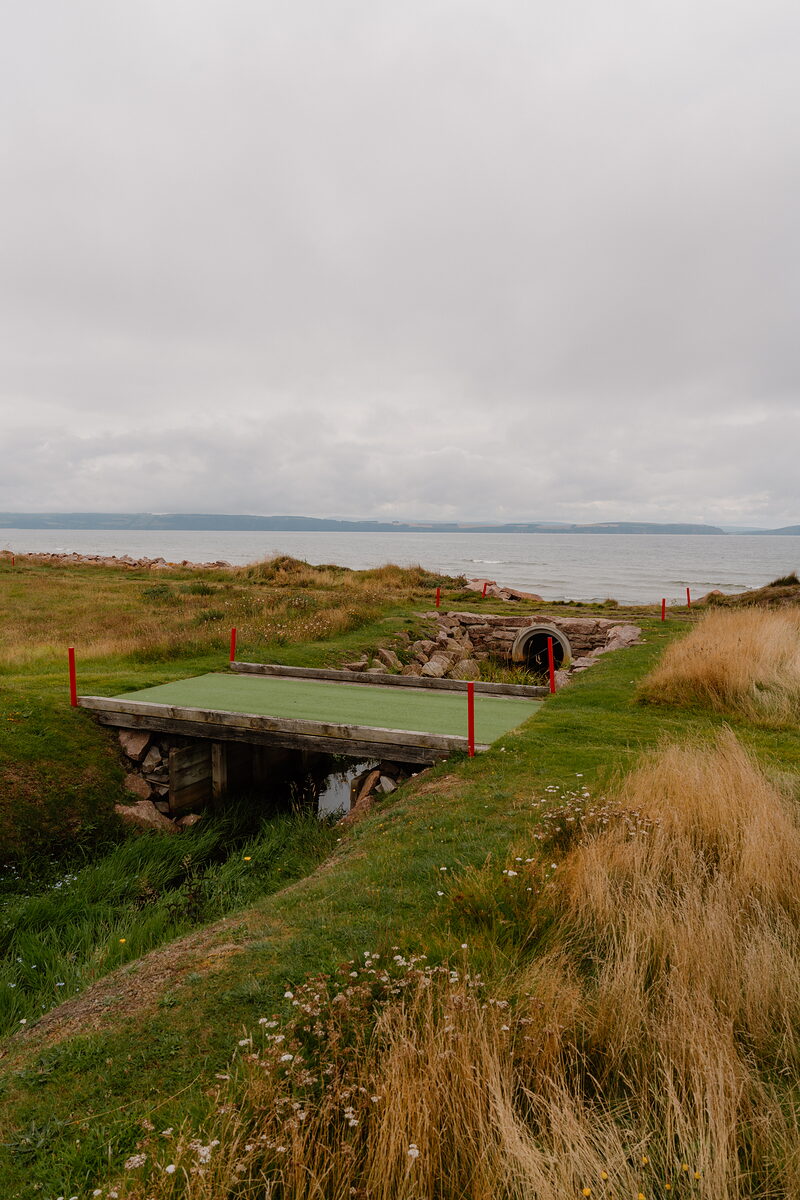 A miniature golf hole crossing a stone culvert, a firth and hills stretching into the distance