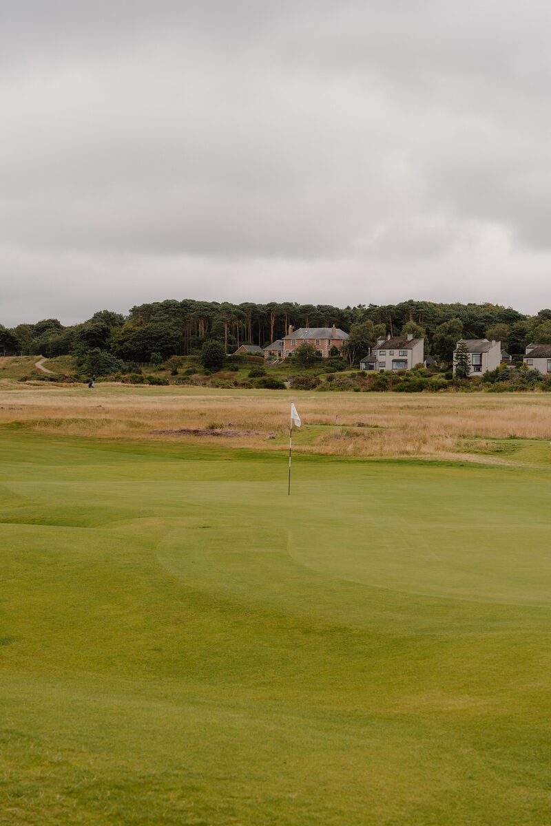 A green marked by a white flag before houses and woodland rising on the horizon