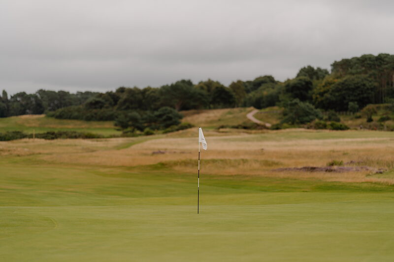 A white flag marking a green bordered by rough grass and distant woodland