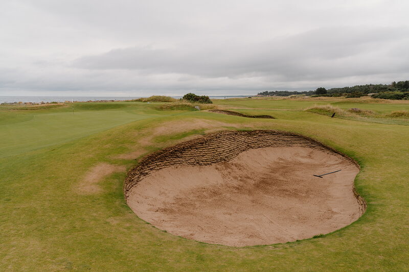 A large bunker beside a fairway bordered by gorse, the sea visible on the horizon