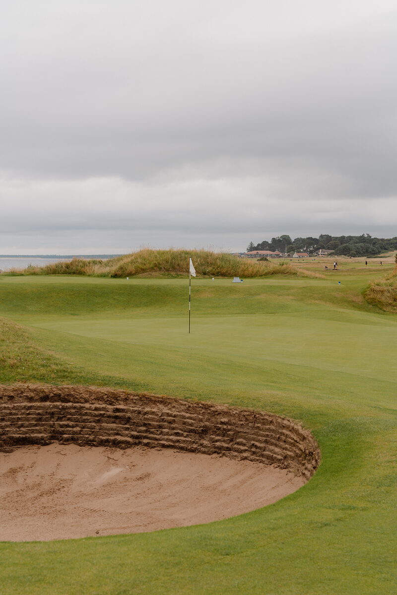 A bunker beside a green marked by a white flag, distant golfers visible on the fairway beyond