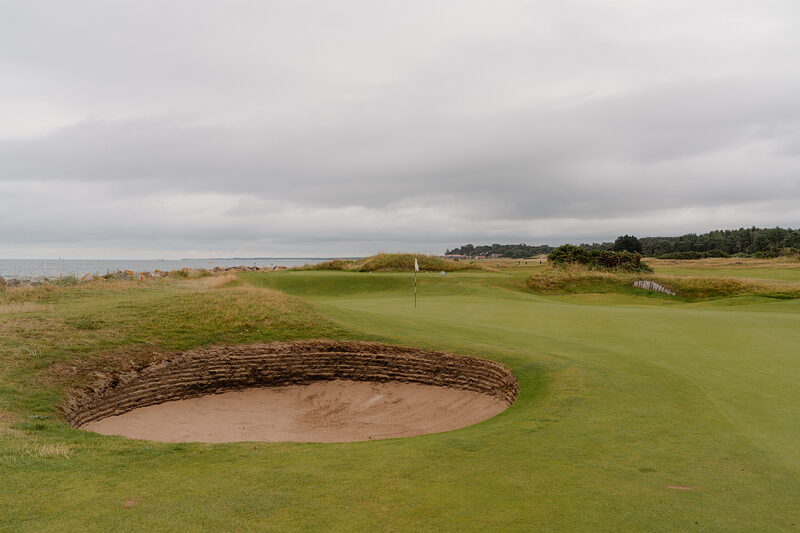 A bunker beside a green marked by a white flag, a firth stretching along the horizon