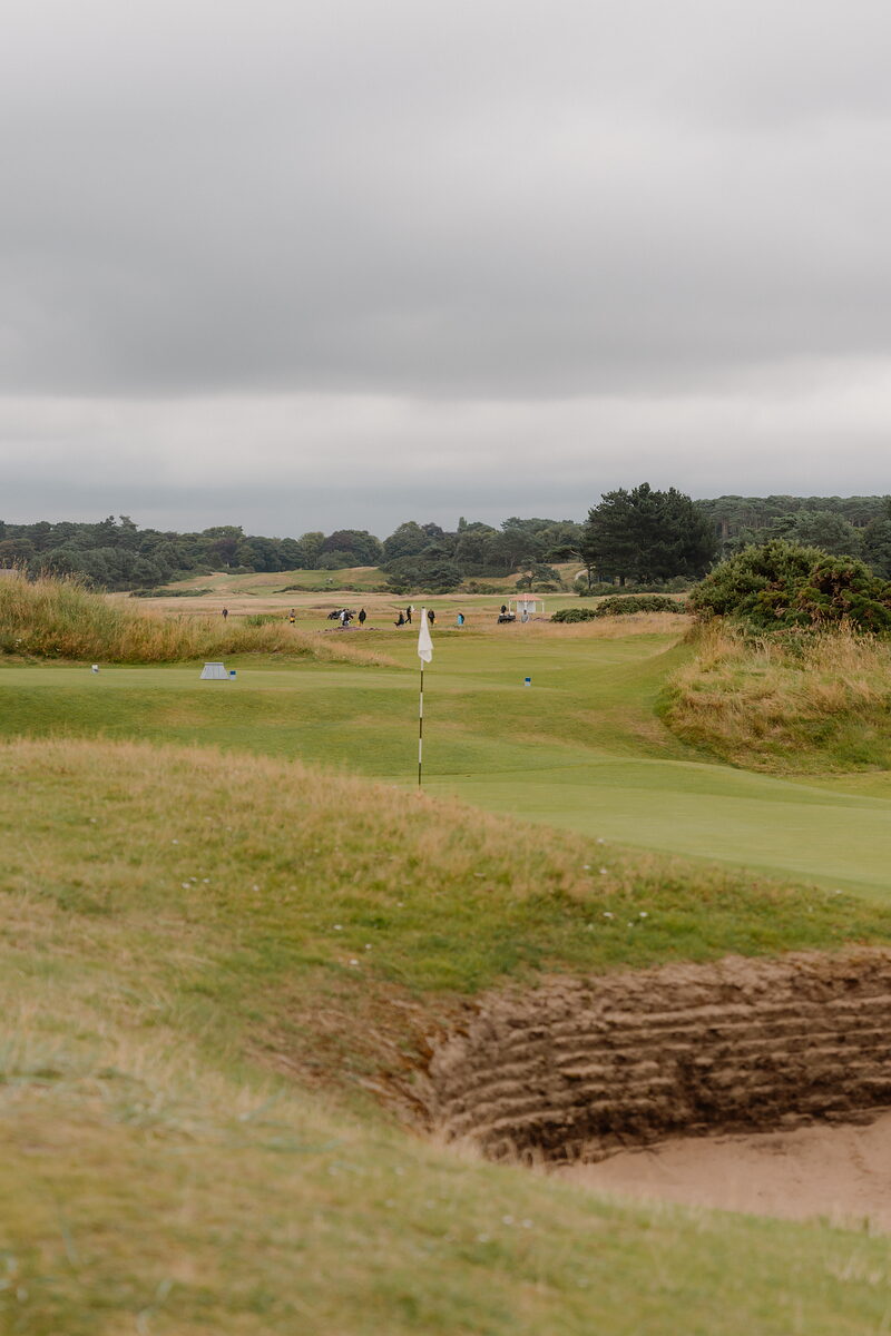 A bunker beside a green marked by a white flag, golfers scattered across the fairway beyond
