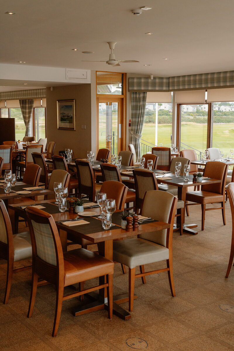 A dining room with tables set in tartan place mats beneath roman blinds overlooking the fairway