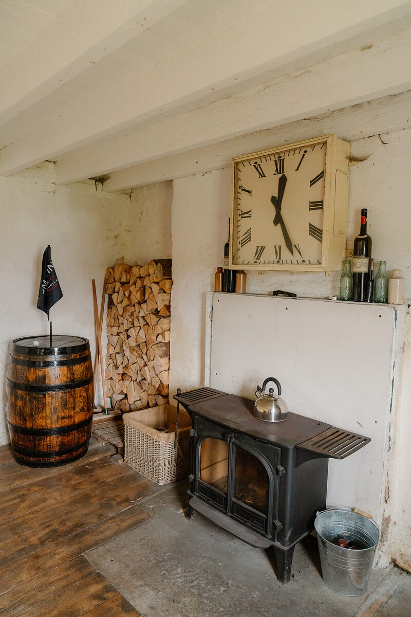 A wood-burning stove beside a whisky barrel and stacked firewood, a square wall clock hanging above