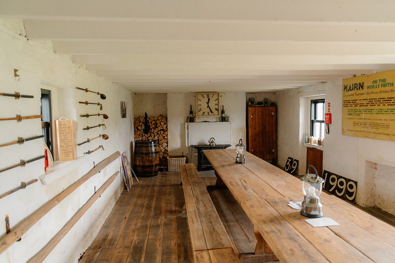 A dining room with wooden benches and a whisky barrel beneath a square wall clock and stacked firewood