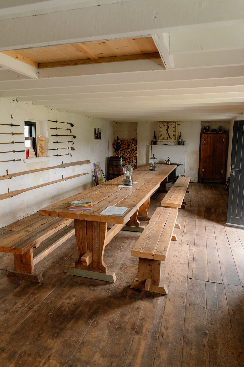 A long wooden table and benches inside a golf clubhouse hall, with clubs mounted on the wall and a wood-burning stove behind