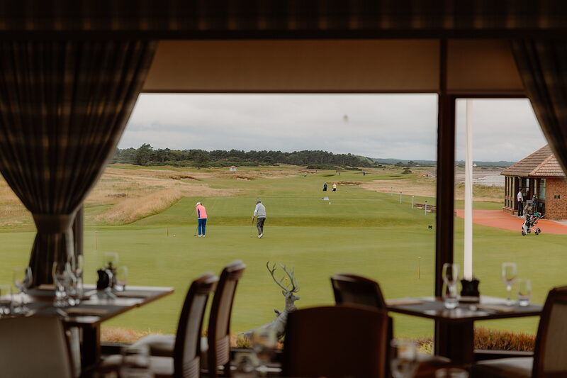 Golfers putting on the green at Nairn, viewed from inside the clubhouse dining room past set tables