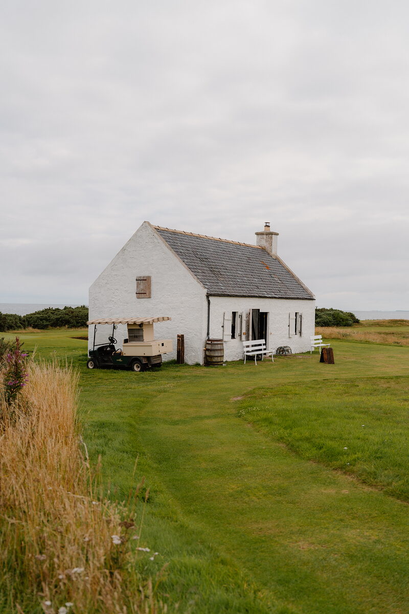 A white-harled cottage with a buggy parked outside, set in open grassland by the course at Nairn