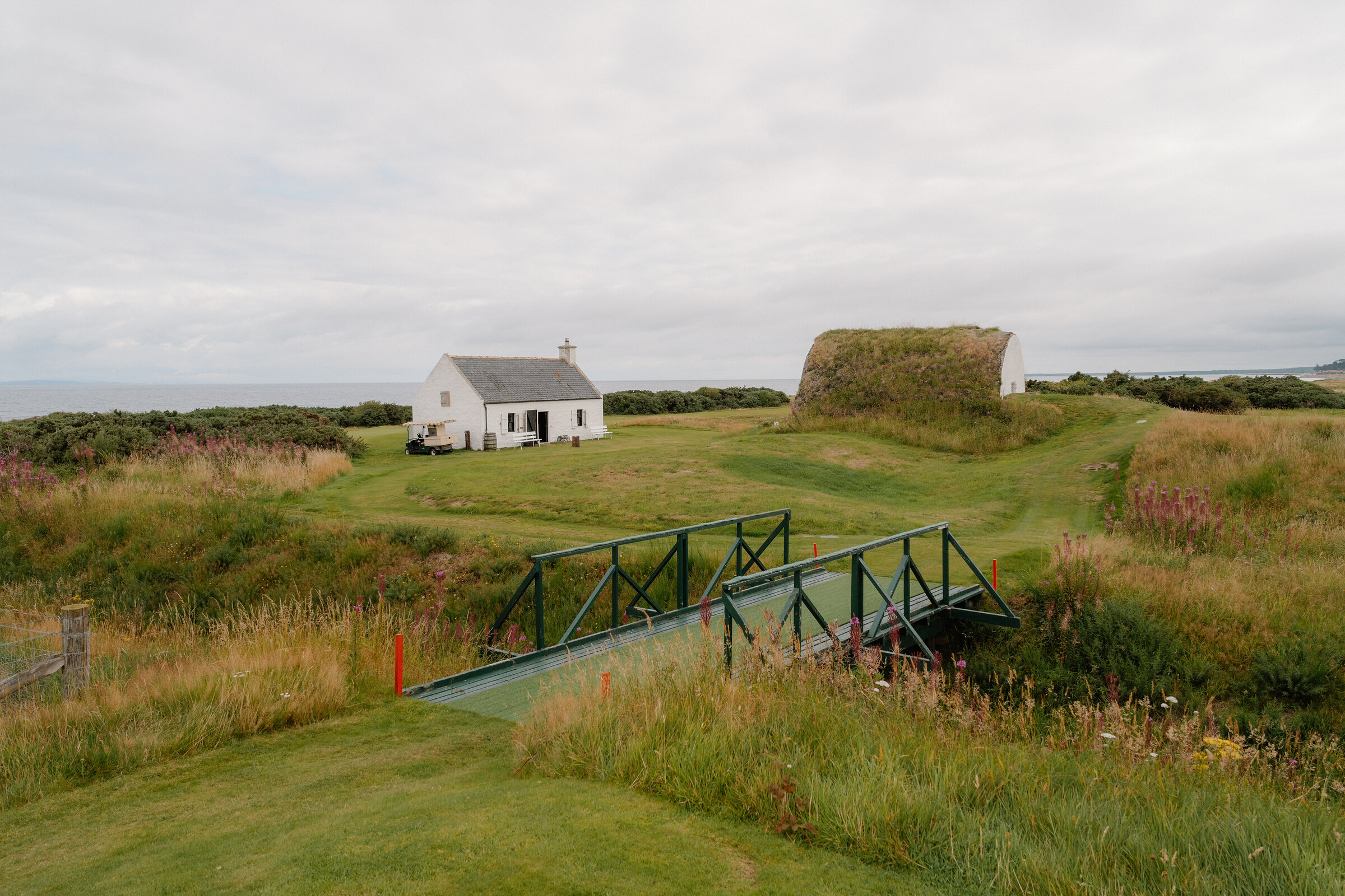 A small white chapel beside a green footbridge over a burn, with an old fort mound and the sea beyond