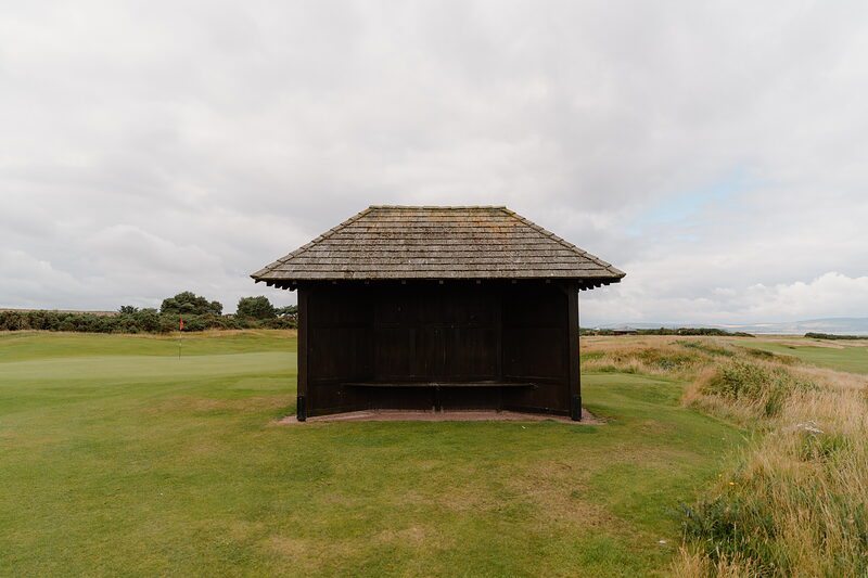 A dark timber shelter with an open front, standing beside the fairway