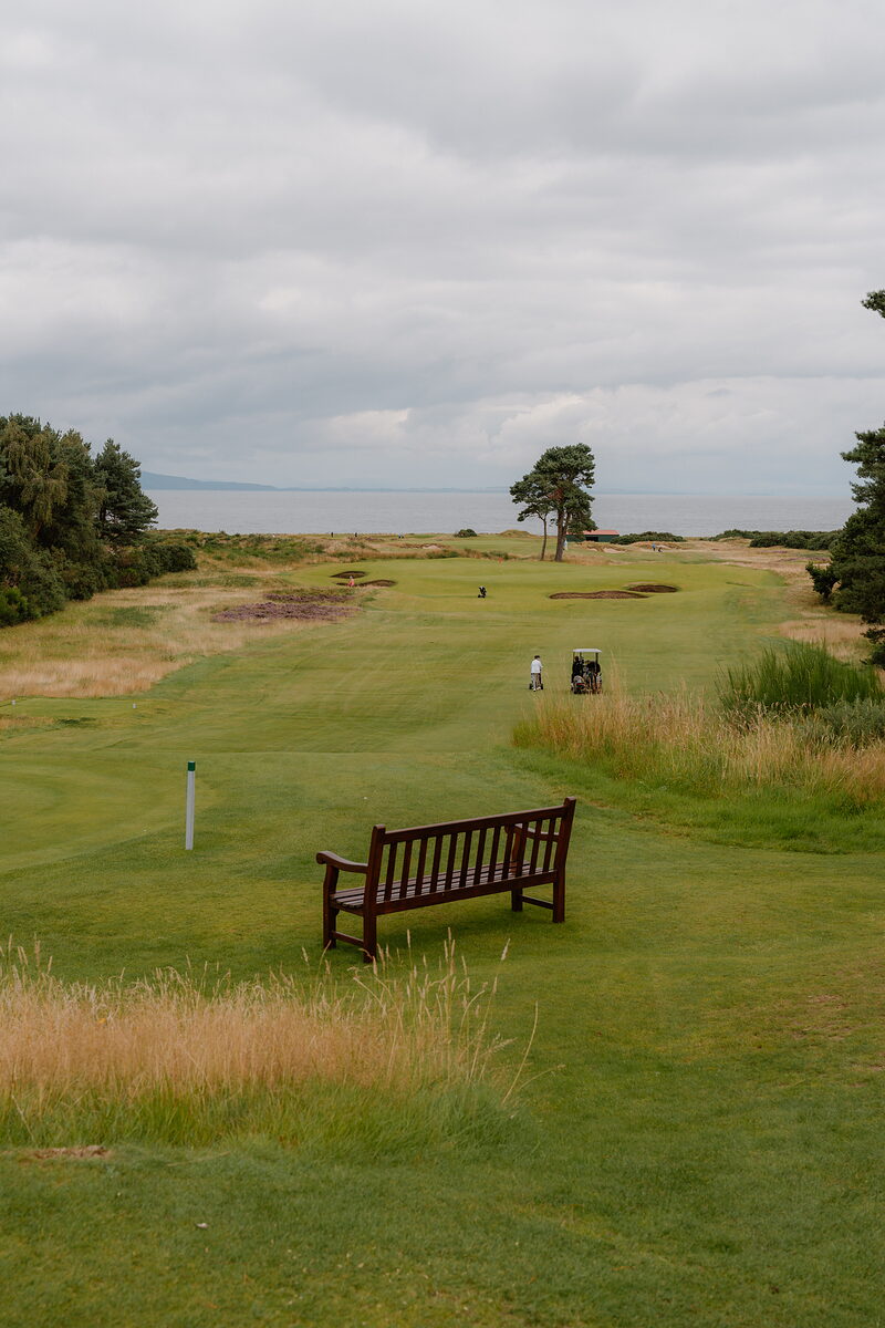 A golfer and buggy on the fairway at Nairn, a wooden bench in the foreground and the sea beyond