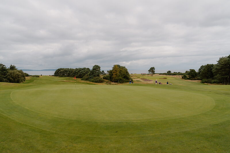 Golfers on the green at Nairn, trees and the sea visible beyond under an overcast sky