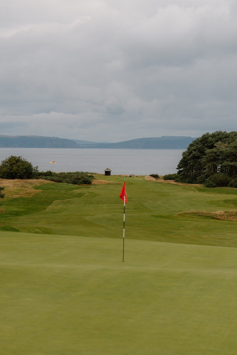 A red flag marking the hole on a links green at Nairn, the coastline curving into the distance