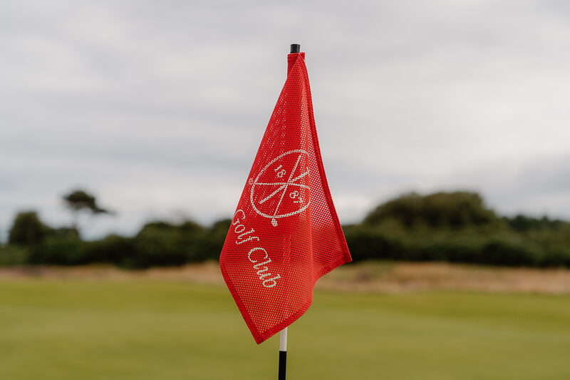 A red flag reading Golf Club marking the hole on a links green, out of focus grass in the foreground